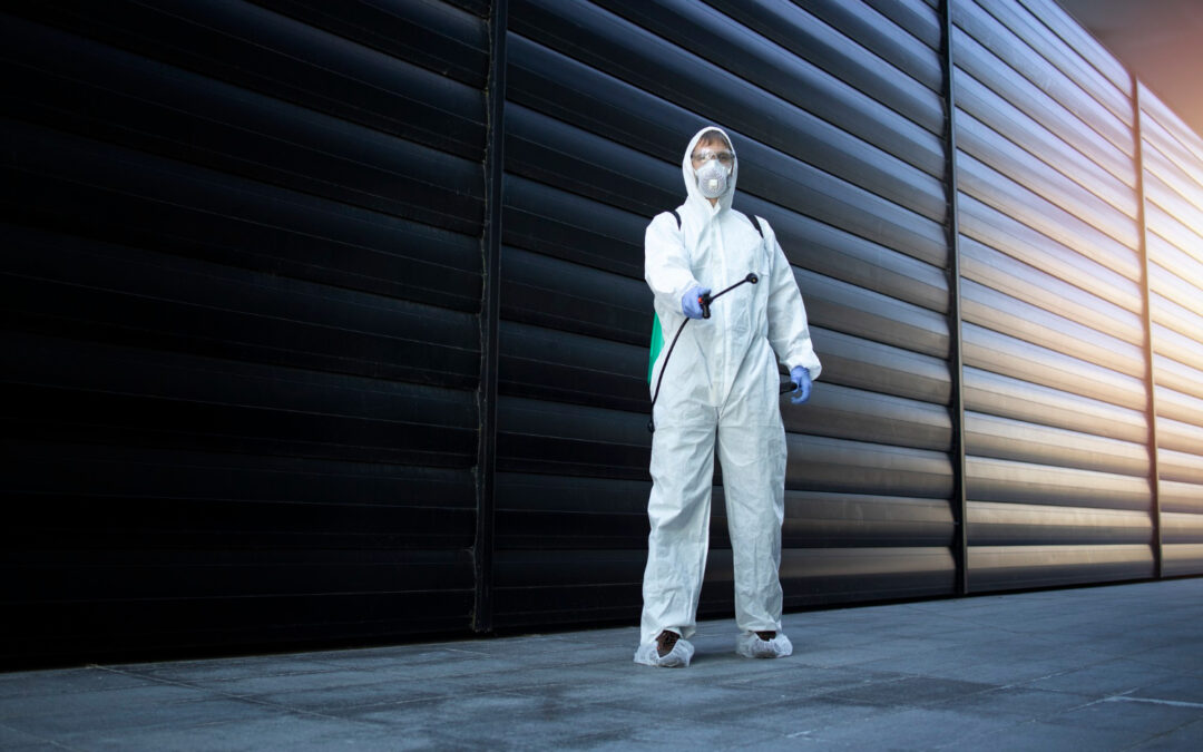 st roberts pest control Person in a white hazmat suit, mask, and gloves holding a sprayer in an industrial or urban setting with a dark wall background.